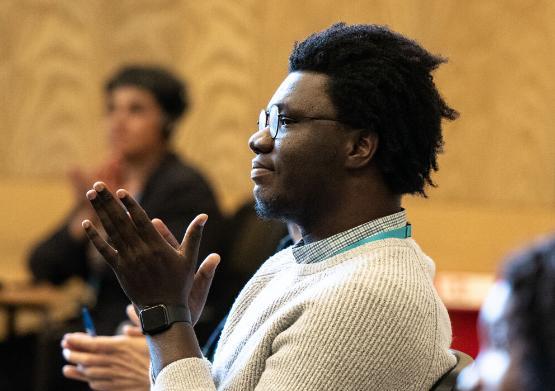 A man with black hair, glasses and a white jumper claps while looking to the left.