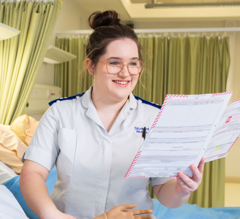 A woman in white uniform stands next to a bed with a training dummy in it.