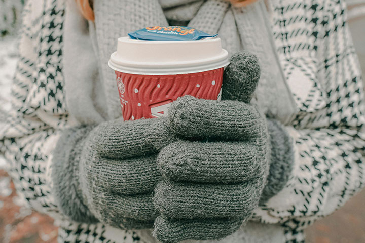 a person wearing a thick pair of grey gloves holds a Costa Coffee branded paper cup. Person is also wearing a scarf and overcoat