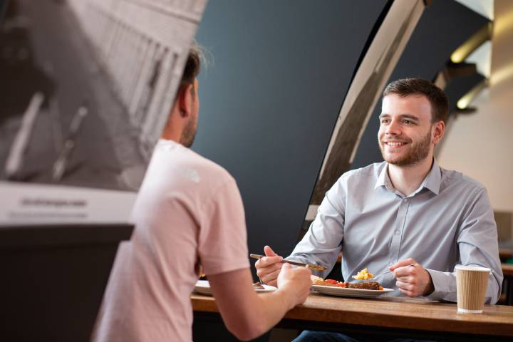 A man sat at a table eating plated food and smiling while sat opposite someone with their back to the camera.