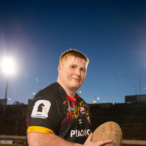 A student dressed in rugby gear standing on the rugby pitch, smiling at the camera while holding a rugby ball