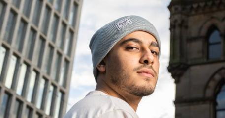 A man in a grey hat stands in front of tall buildings.