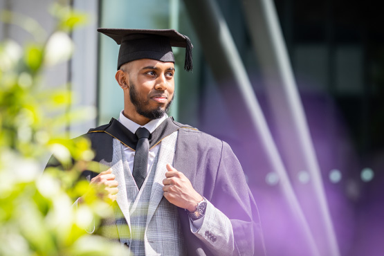 A student posing outside of Richmond Building in a suit and graduation gown and cap.