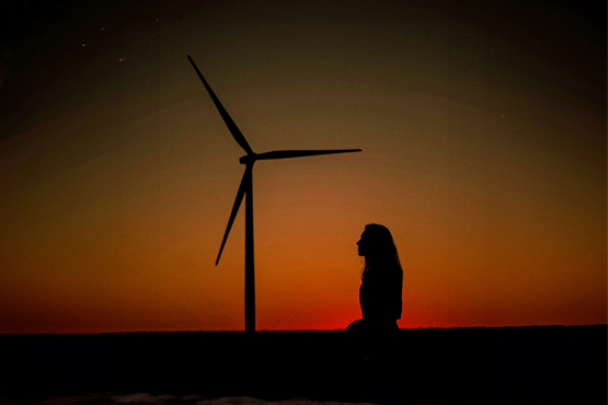 A silhouetted woman kneeling in front of a wind turbine at sunset.