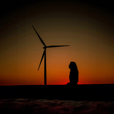 A silhouetted woman kneeling in front of a wind turbine at sunset.