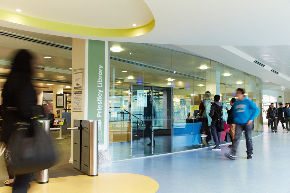 Students walking past the library entrance, with some students looking into the library through a glass window.