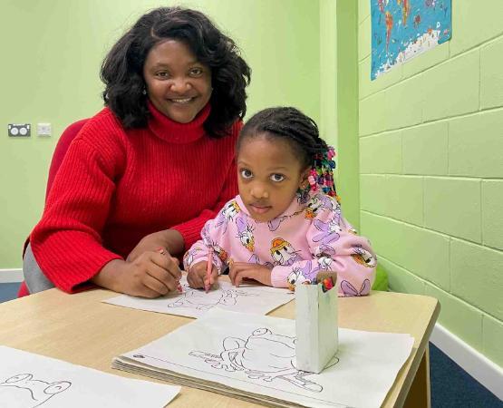 Student sat at small table with young child who is colouring in a picture