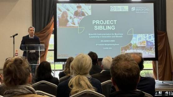 University staff member stood behind podium next to giant screen giving presentation at a conference