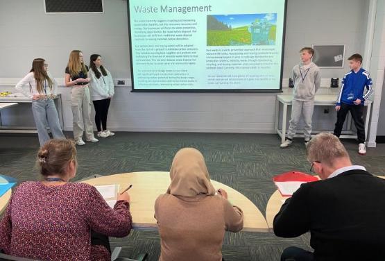 Five students stood either side of a projectrion screen give a pitch to a group of judges sat at a table