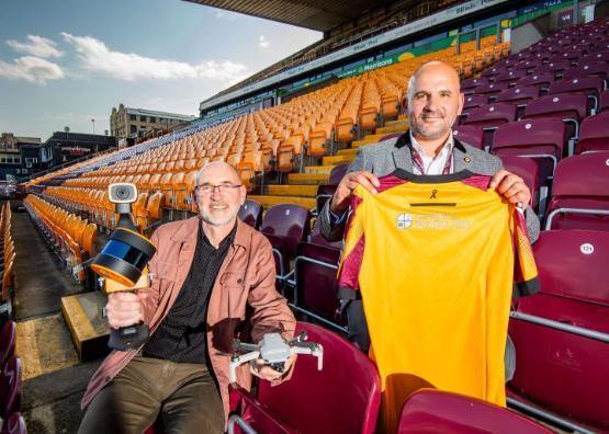 Two people sit next to each other in an empty football stadium's stand, one of them holds a drone while the other person holds a football shirt in front of them