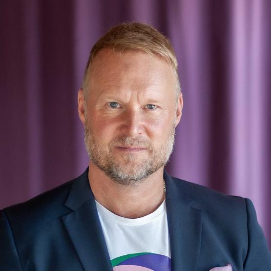 A head and shoulders photograph of a person wearing a T-shirt and jacket poses in front of a purple coloured curtain