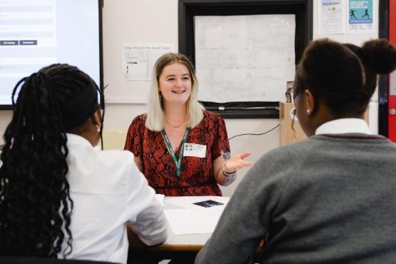 A teacher is sat down in a classroom in front of two school pupils who are sat at another desk in a lesson