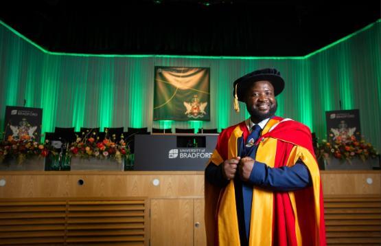 Elgidius Edgar Bwinabona Ichumbaki stands in gown and hat in front of stage at Graduation ceremony