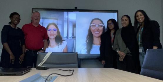 A group of students and academics stand in front of a big screen which has two other students on a video call