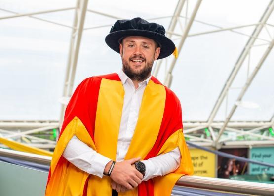 A person in yellow and red graduation robe leans against a glass wall to pose for a photo