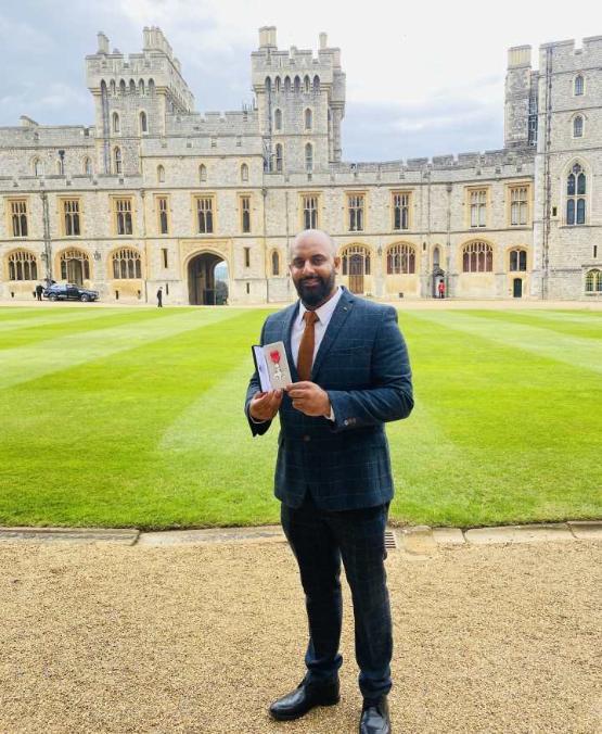 A university staff member dressed in a suit holds up a medal in a box as they stand in the grounds of a castle