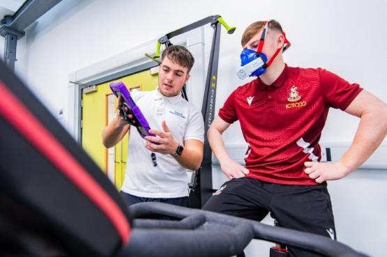 A student tells a football player wearing a mask on an exercise bike about the equipment they are on