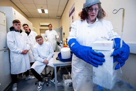 Four people in the background wearing white lab coats look on as a scientist at the front conducts an experiement