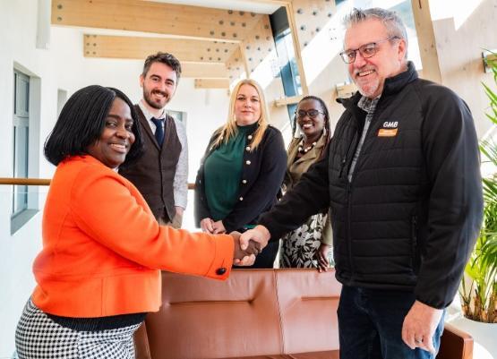 Three people stood up behind two other people at the front who are also stood up shaking hands with each other