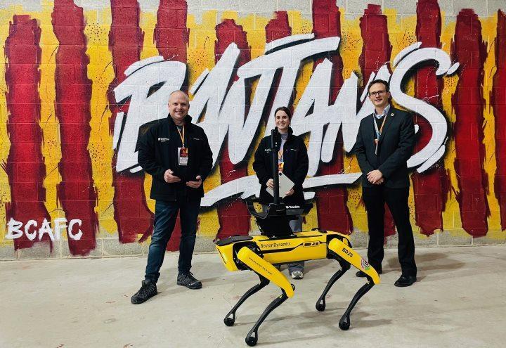 Three university staff members stand in front of claret and amber striped wall art with Bantams written on it with robot dog