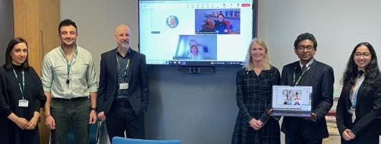 University staff members stood up separated by a video screen behind them which has two people on that screen as part of a video call