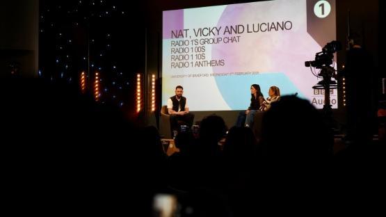 Three people on stage at an event and the photo is taken from the back of the hall which is dimly lit