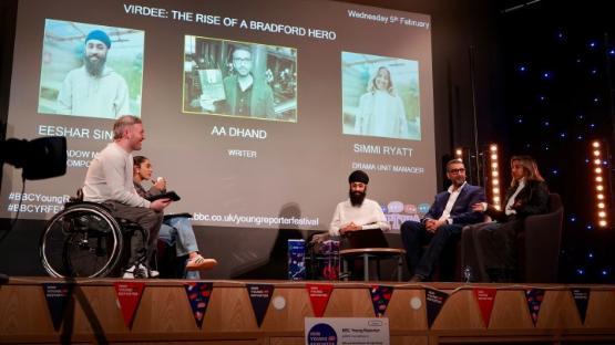 Four people sat on a stage in a discussion at an event with a large screen behind them