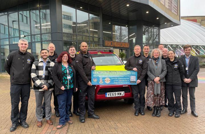 A large group of people stand either wide of a marked fire service vehicle with two of them holding a charity appeal banner