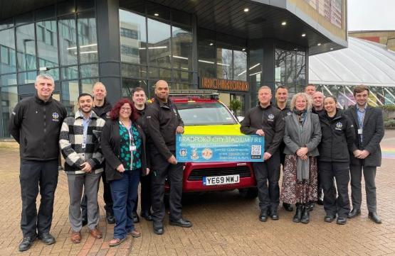 A large group of people stand either wide of a marked fire service vehicle with two of them holding a charity appeal banner