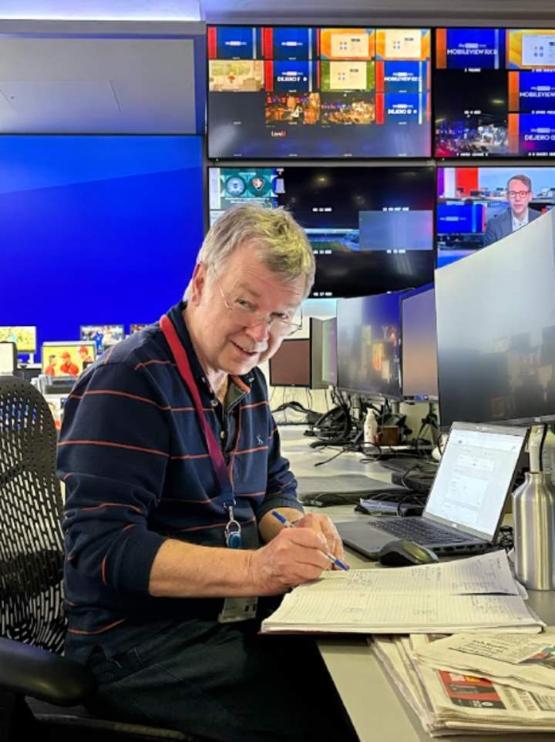 A TV presenter sat in an office with a pen in their right hand writing on paper