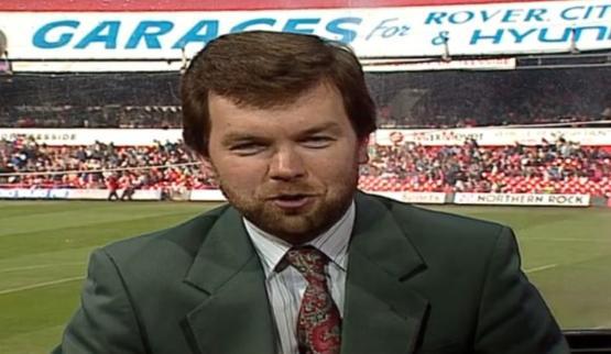 A close-up photo of a TV presenter dressed in a suit with a football stadium behind them