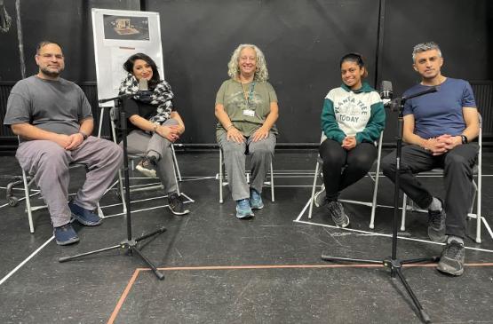 Five people sat on chairs in a play rehearsal room with two large microphones in front of them