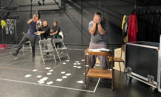 A play rehearsal picture of a character stood up with their hands covering their face while three others are doing a dance and taunting them