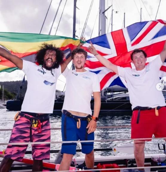 Three graduates dressed in T-shirt and shorts celebrate on a boat holding the United Kingdom and Zimbabwe flags behind them