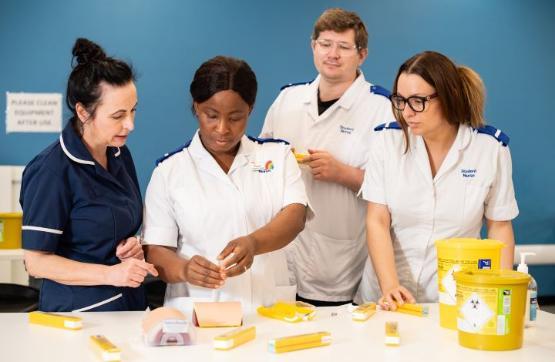 Three nurses in uniform receive training from another nurse in uniform on how to use a drug which is on a table in front of them all