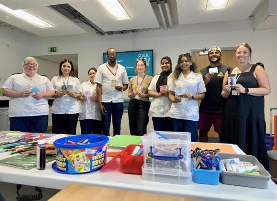 A large group of people, some dressed in nursing uniform, behind a table which has arts and crafts and a sweet box on it