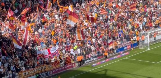 A large crowd of people sat in the stand at a football match with some waving flags