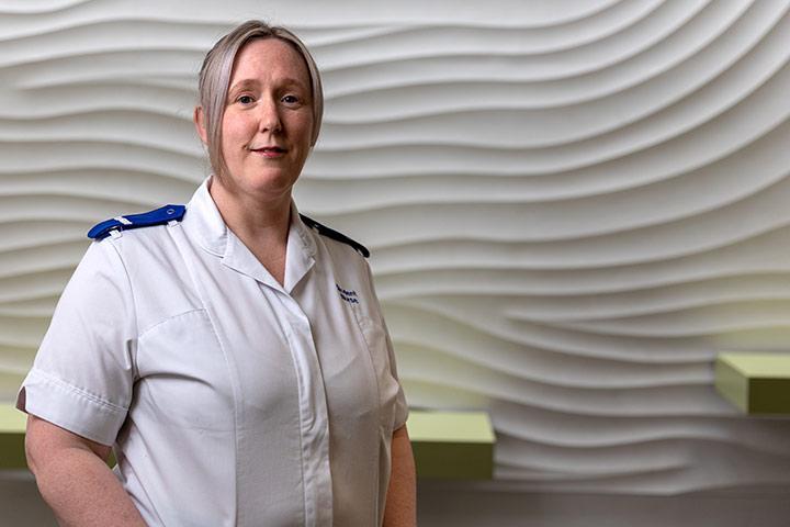 A student in a nursing uniform stood in front of a wave textured background. They are smiling at the camera.