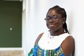 A person in a well-lit indoor space, leaning against a wall and smiling at the camera.