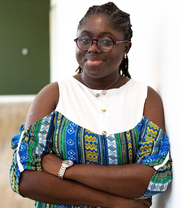 A person in a well lit indoor space, leaning against a wall and smiling at the camera.