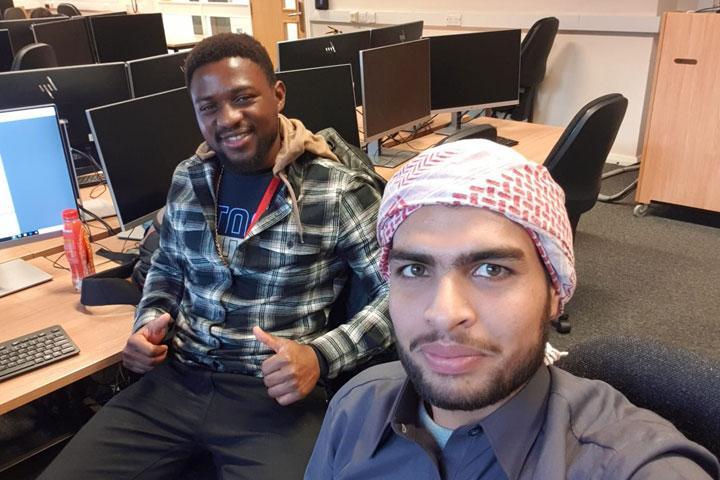 Two students sitting together in a computer lab with computers in the background.