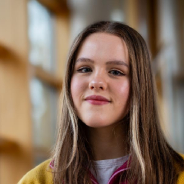 A student with long hair smiles at the camera with large windows in the background.