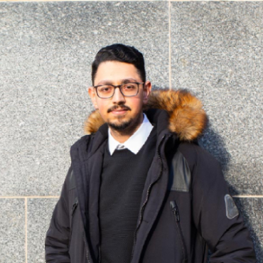 A student with dark hair and a parka looks into the camera with their back to a grey wall.