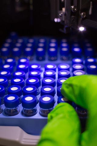 Green glove handling a blue vial in front of a rack of blue vials