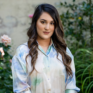 A person facing the camera, standing outside with plants and a building in the background.