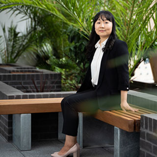 A smartly dressed person sitting on a bench and facing the camera, with large plants in the background.