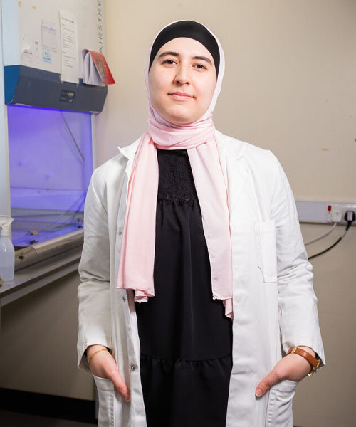 Student wearing a lab coat standing in a lab smiling towards the camera.