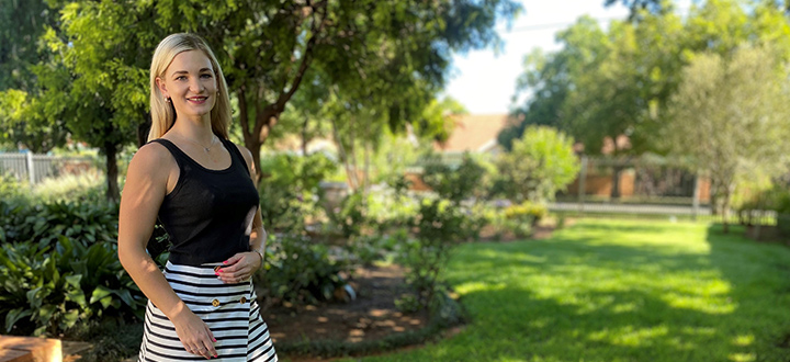 Student standing in front of a sunny luscious green garden while looking at camera and smiling