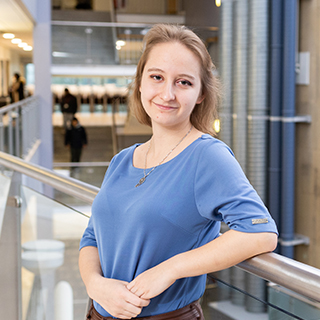 A student wearing a blue top smiles at the camera while leaning against the balcony in the Atrium on the University campus.