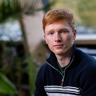 A student smiles as they sit perched on a raised bench infront of indoor plants 